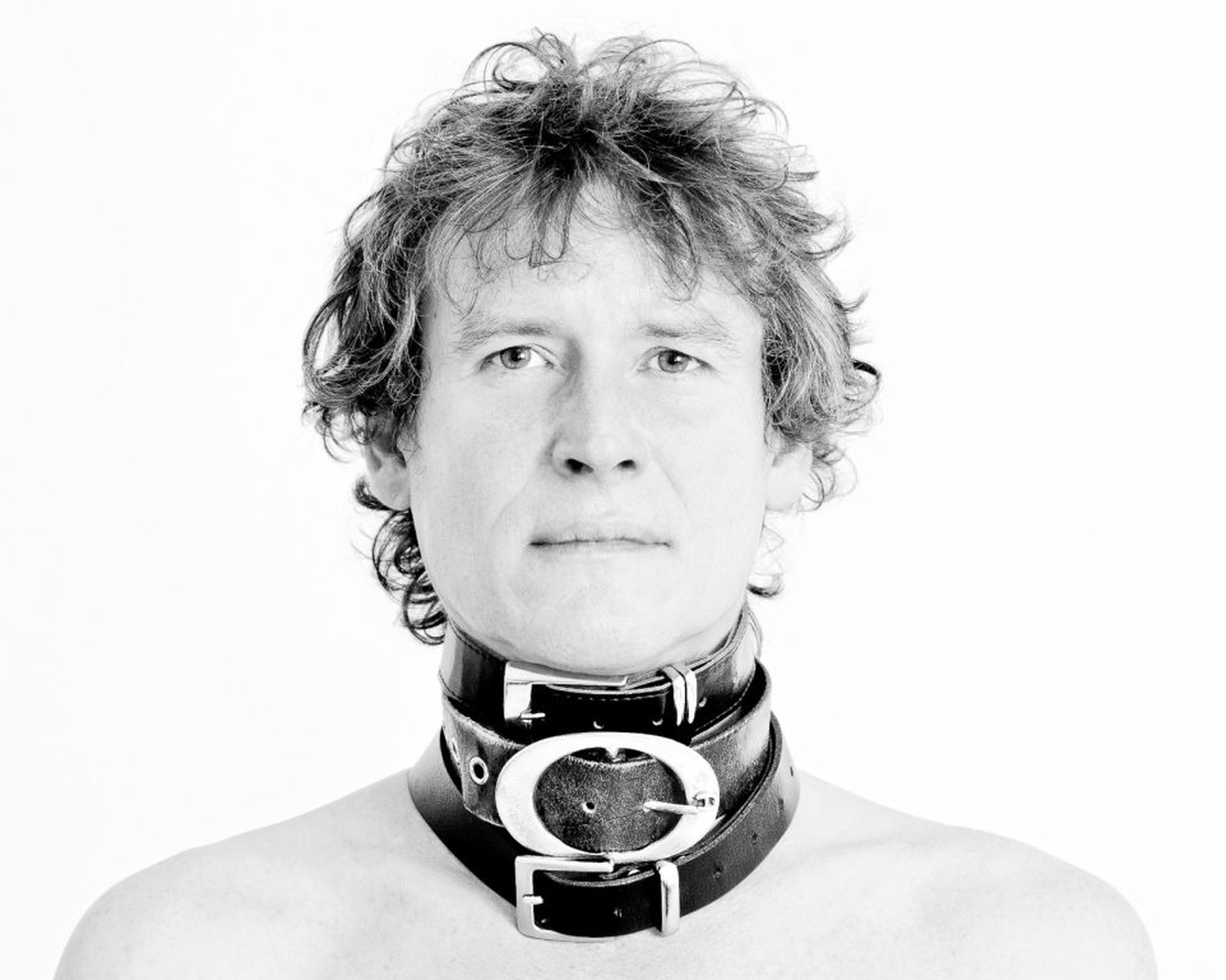 Eine Person mit zerzaustem Haar, die drei übereinanderliegende schwarze Ledergürtel um den Hals trägt. (KI generierte Beschreibung)