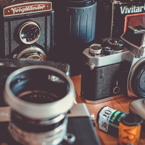 Auf einem Tisch angeordnete Vintage-Kameras und Fotoausrüstung, darunter eine Nikon-Spiegelreflexkamera, eine Voigtländer und Filmrollen. (KI generierte Beschreibung)
