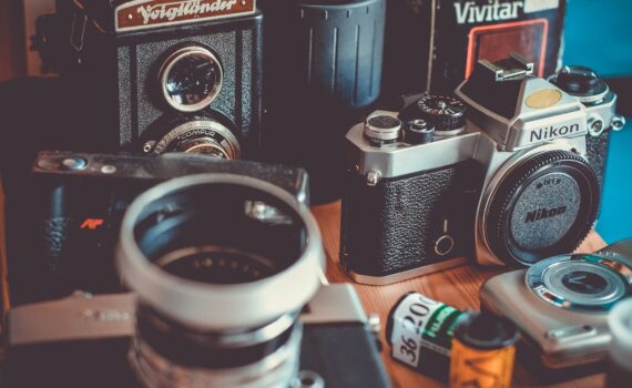 Auf einem Tisch angeordnete Vintage-Kameras und Fotoausrüstung, darunter eine Nikon-Spiegelreflexkamera, eine Voigtländer und Filmrollen. (KI generierte Beschreibung)