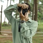 Eine Person in einem hellgrünen Hemd hält eine Kamera an ihr Gesicht. Sie steht im Freien in einem Waldgebiet mit Bäumen und Seilsperren im Hintergrund. (KI generierte Beschreibung)