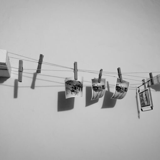Vier kleine Fotos hängen an einer Schnur mit Wäscheklammern, die an zwei Schaumstoffblöcken an einer weißen Wand befestigt sind. Das Bild ist in schwarz-weiß gehalten. (KI generierte Beschreibung)