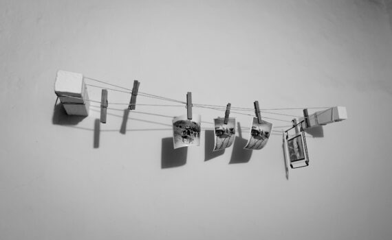 Vier kleine Fotos hängen an einer Schnur mit Wäscheklammern, die an zwei Schaumstoffblöcken an einer weißen Wand befestigt sind. Das Bild ist in schwarz-weiß gehalten. (KI generierte Beschreibung)