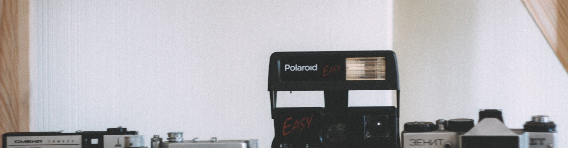 Vier alte Kameras, darunter eine Polaroid und drei Filmkameras, sind auf einem Holzregal vor einem neutralen Hintergrund ausgestellt. (KI generierte Beschreibung)