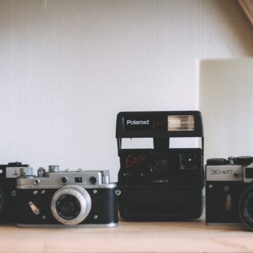 Vier alte Kameras, darunter eine Polaroid und drei Filmkameras, sind auf einem Holzregal vor einem neutralen Hintergrund ausgestellt. (KI generierte Beschreibung)