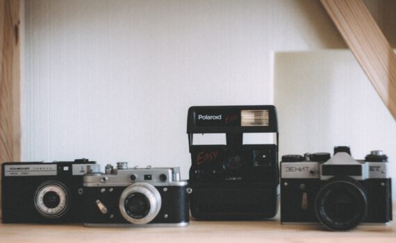 Vier alte Kameras, darunter eine Polaroid und drei Filmkameras, sind auf einem Holzregal vor einem neutralen Hintergrund ausgestellt. (KI generierte Beschreibung)