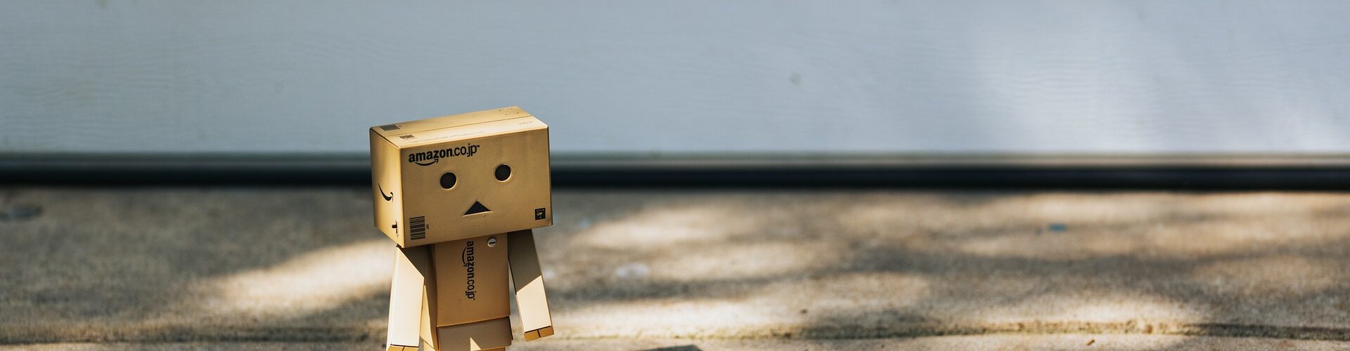 Eine kleine Pappfigur, die einem Roboter ähnelt, steht im Sonnenlicht neben einer schattigen Tür und vermittelt ein Gefühl von Neugier und Entdeckergeist. (KI generierte Beschreibung)