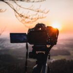 Mit einer Digitalkamera auf einem Stativ wird eine malerische Landschaft bei Sonnenuntergang fotografiert, wobei die Sonne tief am Horizont steht und die Äste darüber sichtbar sind. (KI generierte Beschreibung)