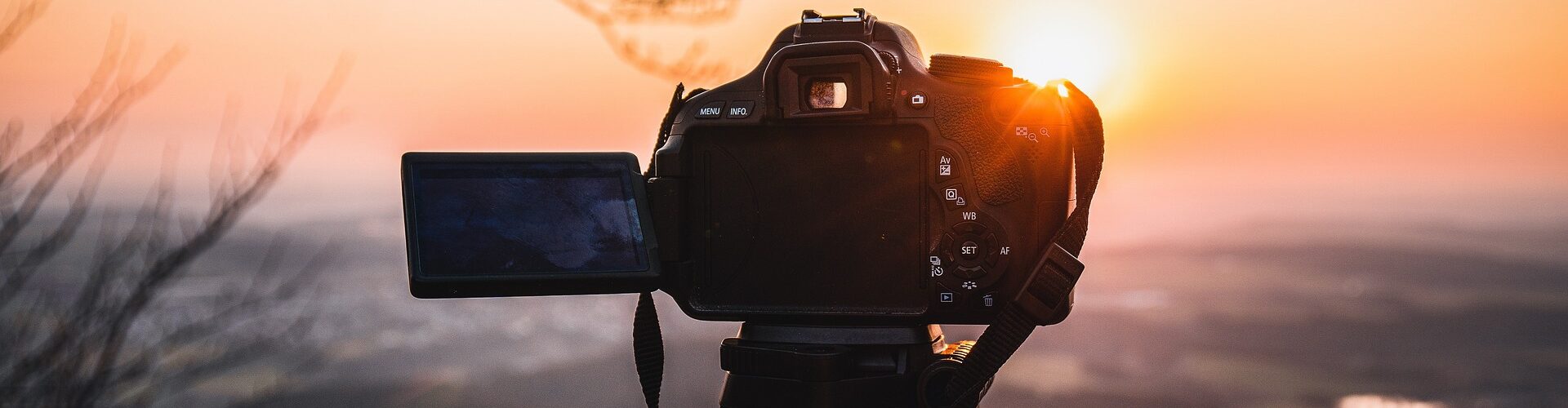 Mit einer Digitalkamera auf einem Stativ wird eine malerische Landschaft bei Sonnenuntergang fotografiert, wobei die Sonne tief am Horizont steht und die Äste darüber sichtbar sind. (KI generierte Beschreibung)