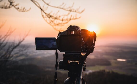 Mit einer Digitalkamera auf einem Stativ wird eine malerische Landschaft bei Sonnenuntergang fotografiert, wobei die Sonne tief am Horizont steht und die Äste darüber sichtbar sind. (KI generierte Beschreibung)