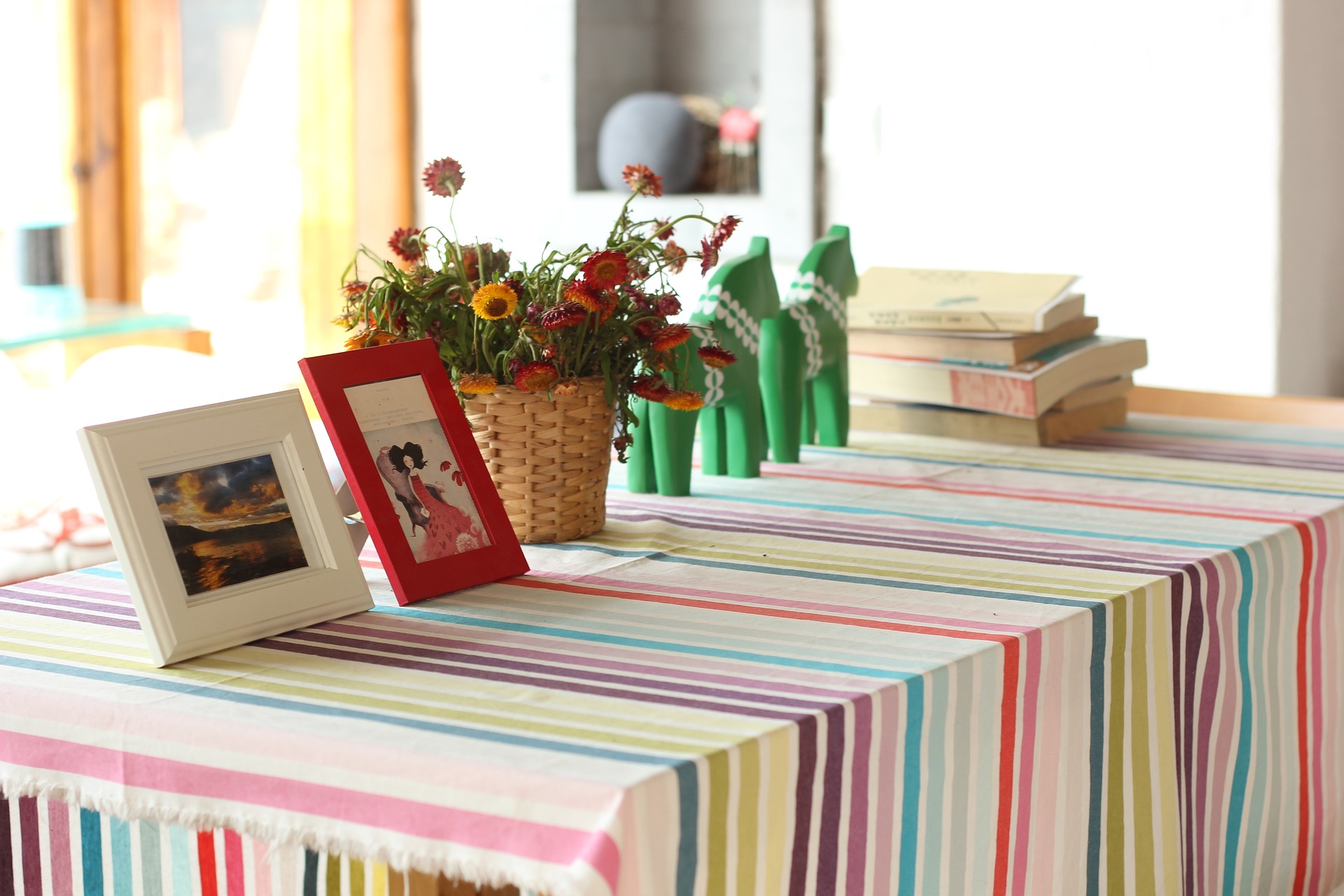 Eine bunt gestreifte Tischdecke, darauf ein Korb mit Wildblumen, gerahmte Bilder und ein Stapel Bücher in einem hellen Raum. . (KI generierte Beschreibung)