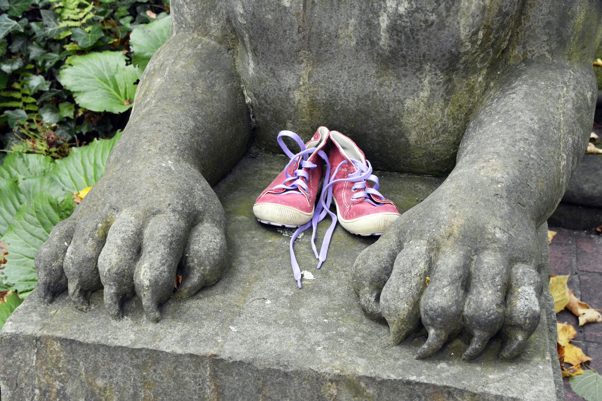 Ein Paar kleine rote Turnschuhe mit offenen Schnürsenkeln liegt auf dem Schoß einer Steinstatue mit großen Pfoten. (KI generierte Beschreibung)