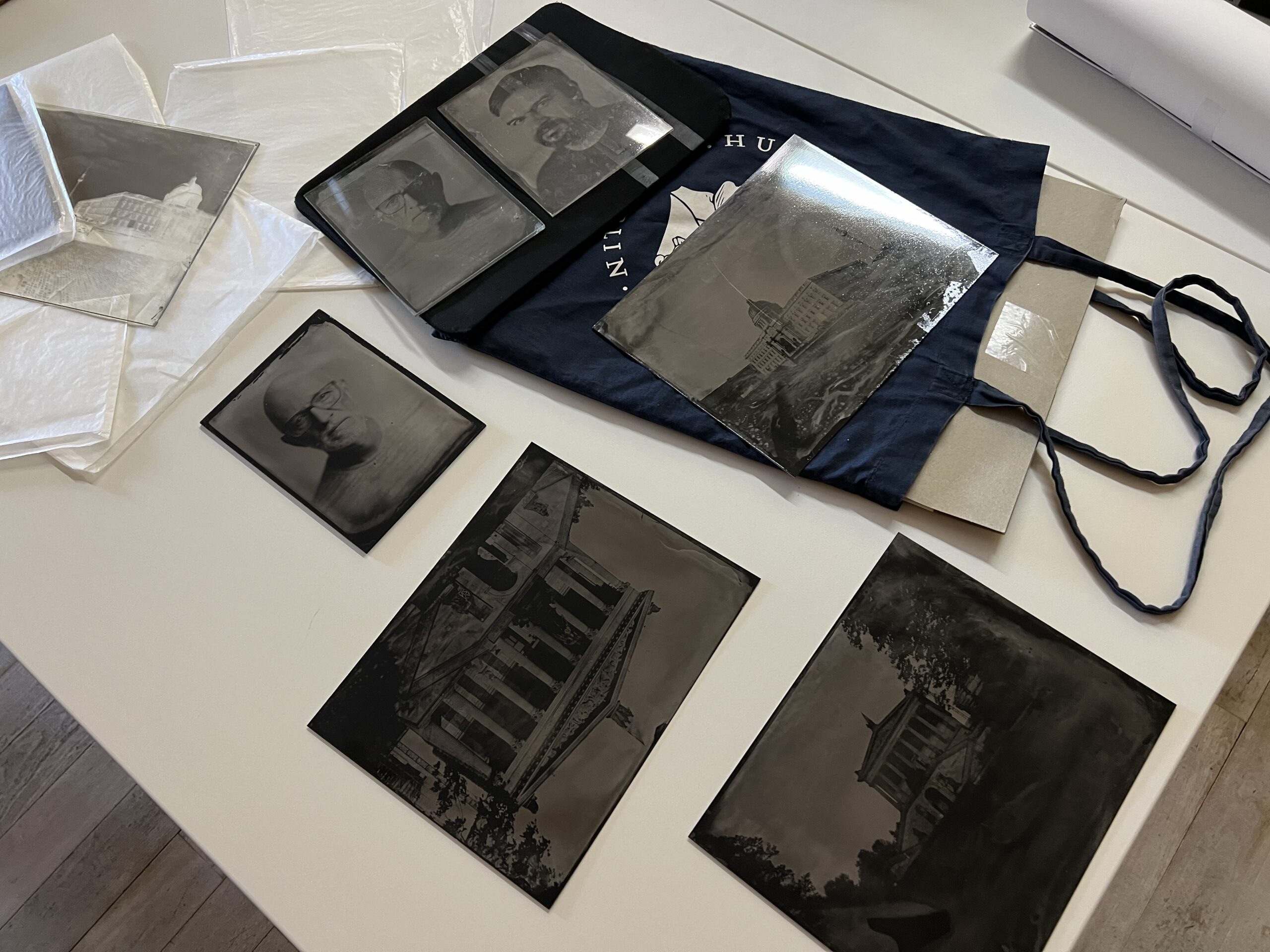Auf einem Tisch sind mehrere schwarz-weiße fotografische Glasnegative von Gebäuden und Porträts ausgestellt, die teilweise in Schutzhüllen und einer Stoffhülle eingelegt sind. (KI generierte Beschreibung)
