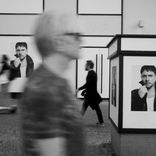 Schwarzweißfoto von Fußgängern, die auf einer Stadtstraße an großen Plakaten eines Mannes vorbeigehen. (KI generierte Beschreibung)