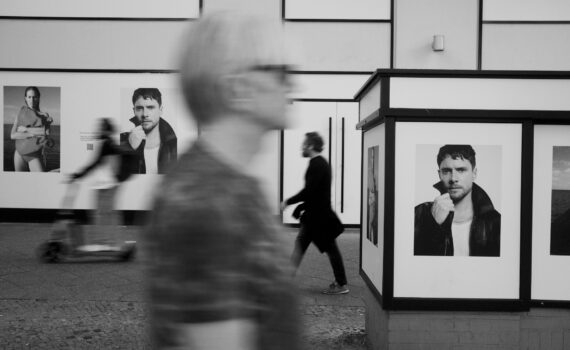 Schwarzweißfoto von Fußgängern, die auf einer Stadtstraße an großen Plakaten eines Mannes vorbeigehen. (KI generierte Beschreibung)