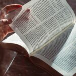 Eine Hand blättert im Tageslicht die Seite eines aufgeschlagenen Buches mit einem gelben Lesezeichenband um. (KI generierte Beschreibung)