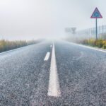 Eine neblige Straße mit sichtbaren Verkehrsschildern an den Seiten und einem Zaun im Hintergrund, die in dichten Nebel führt. (KI generierte Beschreibung)