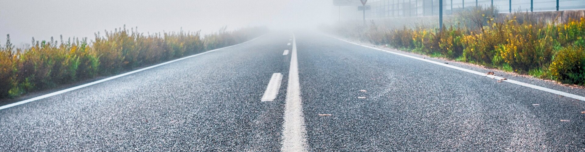 Eine neblige Straße mit sichtbaren Verkehrsschildern an den Seiten und einem Zaun im Hintergrund, die in dichten Nebel führt. (KI generierte Beschreibung)