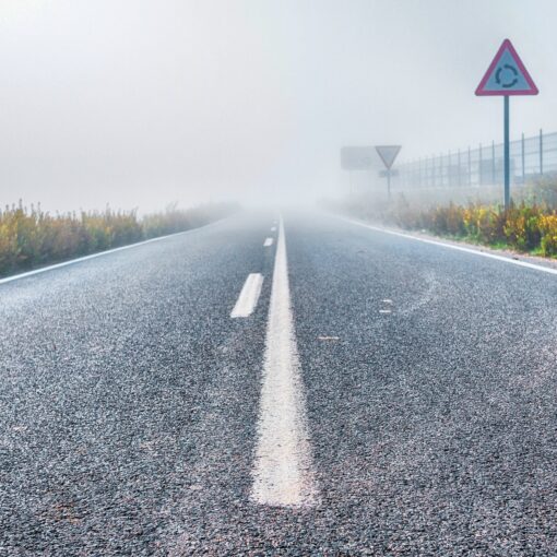 Eine neblige Straße mit sichtbaren Verkehrsschildern an den Seiten und einem Zaun im Hintergrund, die in dichten Nebel führt. (KI generierte Beschreibung)