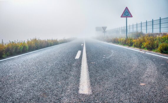 Eine neblige Straße mit sichtbaren Verkehrsschildern an den Seiten und einem Zaun im Hintergrund, die in dichten Nebel führt. (KI generierte Beschreibung)