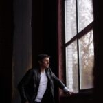Eine Person in einer Lederjacke steht an einem großen Fenster und schaut hinaus. (KI generierte Beschreibung)