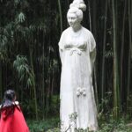 Ein Kind in einem roten Umhang steht vor einer großen Steinstatue einer Frau in einem Bambuswald. (KI generierte Beschreibung)