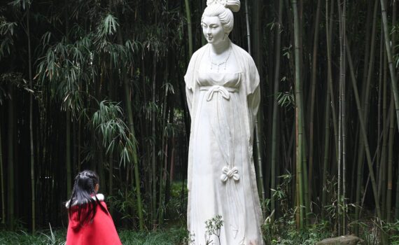 Ein Kind in einem roten Umhang steht vor einer großen Steinstatue einer Frau in einem Bambuswald. (KI generierte Beschreibung)