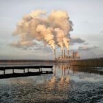 Kraftwerk mit vier Schornsteinen, deren Emissionen in den Himmel strömen, der sich in einem nahe gelegenen Gewässer mit einem Dock im Vordergrund spiegelt. (KI generierte Beschreibung)