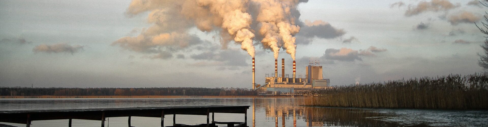 Kraftwerk mit vier Schornsteinen, deren Emissionen in den Himmel strömen, der sich in einem nahe gelegenen Gewässer mit einem Dock im Vordergrund spiegelt. (KI generierte Beschreibung)