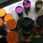 Auf einer Metalloberfläche sind Farbdosen angeordnet, einige davon offen, andere geschlossen. Zu den sichtbaren Farbtönen gehören Orange, Lila und Grün. (KI generierte Beschreibung)