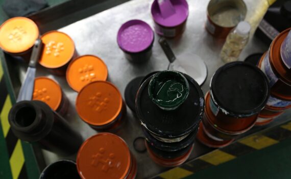 Auf einer Metalloberfläche sind Farbdosen angeordnet, einige davon offen, andere geschlossen. Zu den sichtbaren Farbtönen gehören Orange, Lila und Grün. (KI generierte Beschreibung)