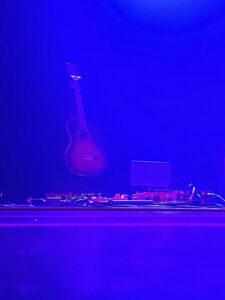 An einer Wand hängt eine Gitarre, auf dem Boden liegen Audiogeräte und Kabel, angestrahlt von blauer Bühnenbeleuchtung. (KI generierte Beschreibung)