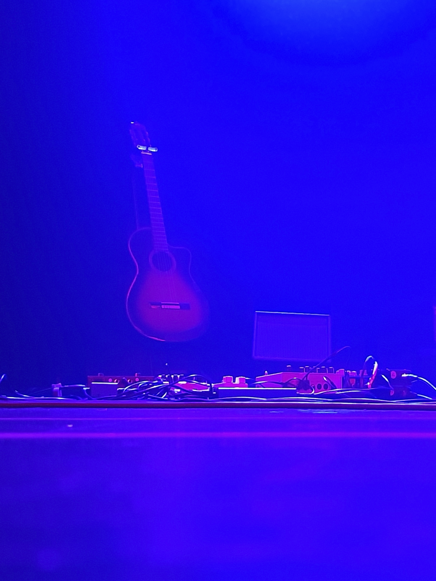 An einer Wand hängt eine Gitarre, auf dem Boden liegen Audiogeräte und Kabel, angestrahlt von blauer Bühnenbeleuchtung. (KI generierte Beschreibung)