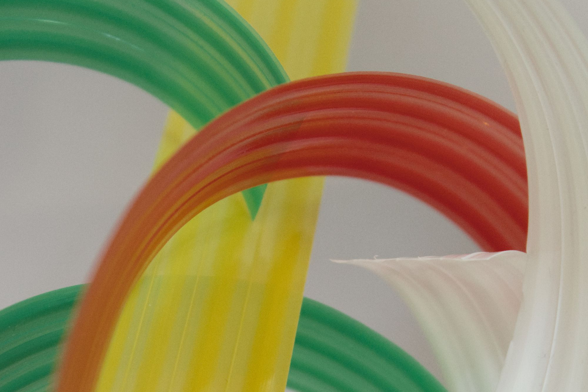 Farbenfrohe, gebogene Glasskulpturen mit grünen, roten, gelben und weißen Bögen, die vor einem neutralen Hintergrund ineinander verschlungen sind. (KI generierte Beschreibung)