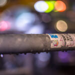 Vor einem verschwommenen, bunten Hintergrund aus Bokeh-Lichtern ist ein nasser Metallpfosten mit abblätternden Aufklebern im Fokus. (KI generierte Beschreibung)