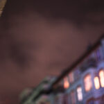 Eine Nachtszene mit verschwommenen Gebäuden und leuchtenden Fenstern unter einem bewölkten Himmel. Im Vordergrund ist ein tropfender Wasserhahn zu sehen. (KI generierte Beschreibung)