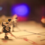 Nahaufnahme eines Tischhockeyspiels. Zu sehen sind kleine Hockeyspieler aus Plastik auf einer Eisfläche mit verschwommenem violettem Licht im Hintergrund. (KI generierte Beschreibung)