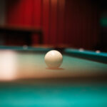 Nahaufnahme einer weißen Billardkugel auf einem grünen Billardtisch mit einem unscharfen Hintergrund in Rot- und Dunkeltönen. (KI generierte Beschreibung)