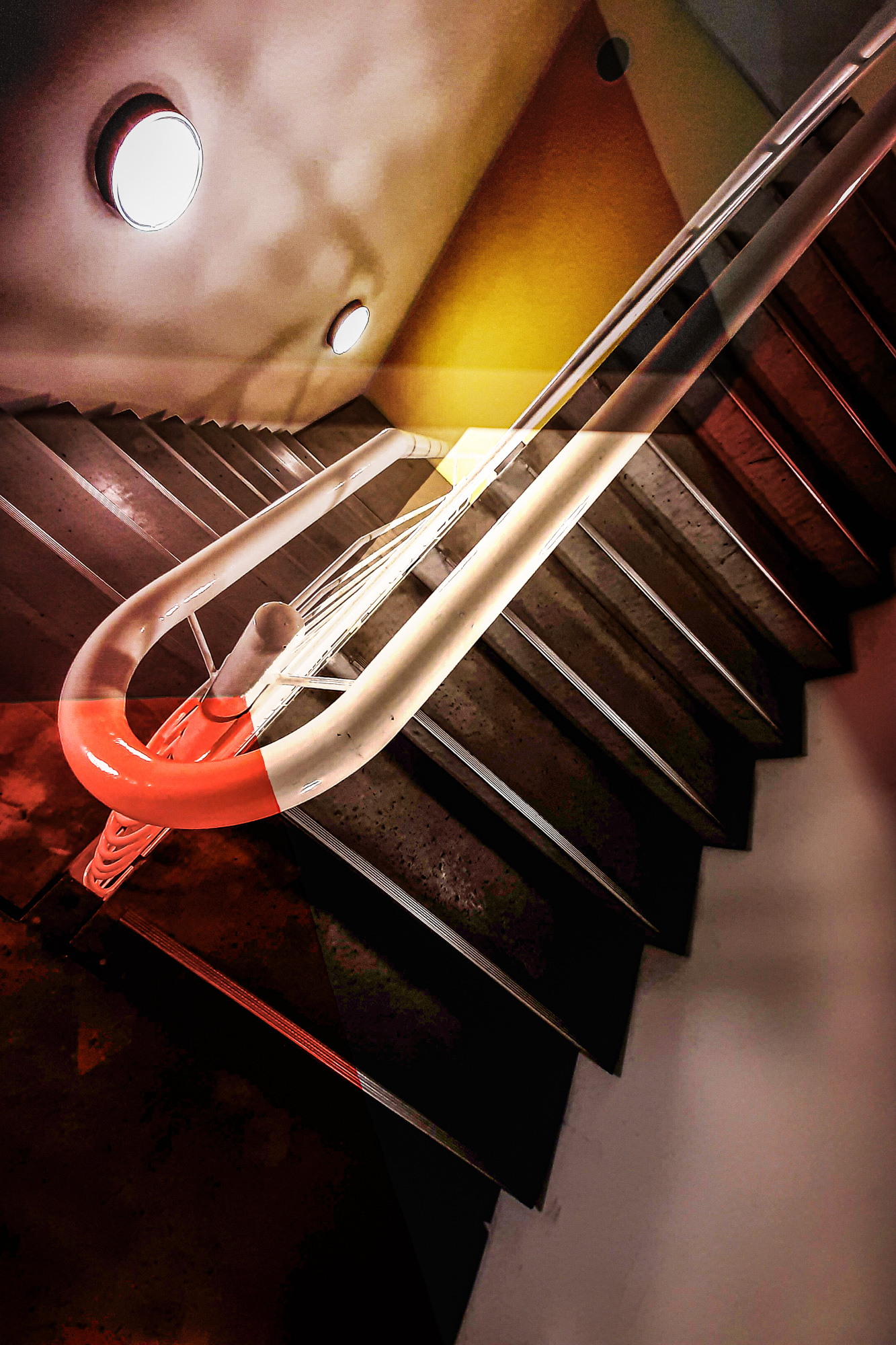 Eine Wendeltreppe mit Metallgeländer, beleuchtet durch Deckenleuchten, aus einem Winkel betrachtet. (KI generierte Beschreibung)