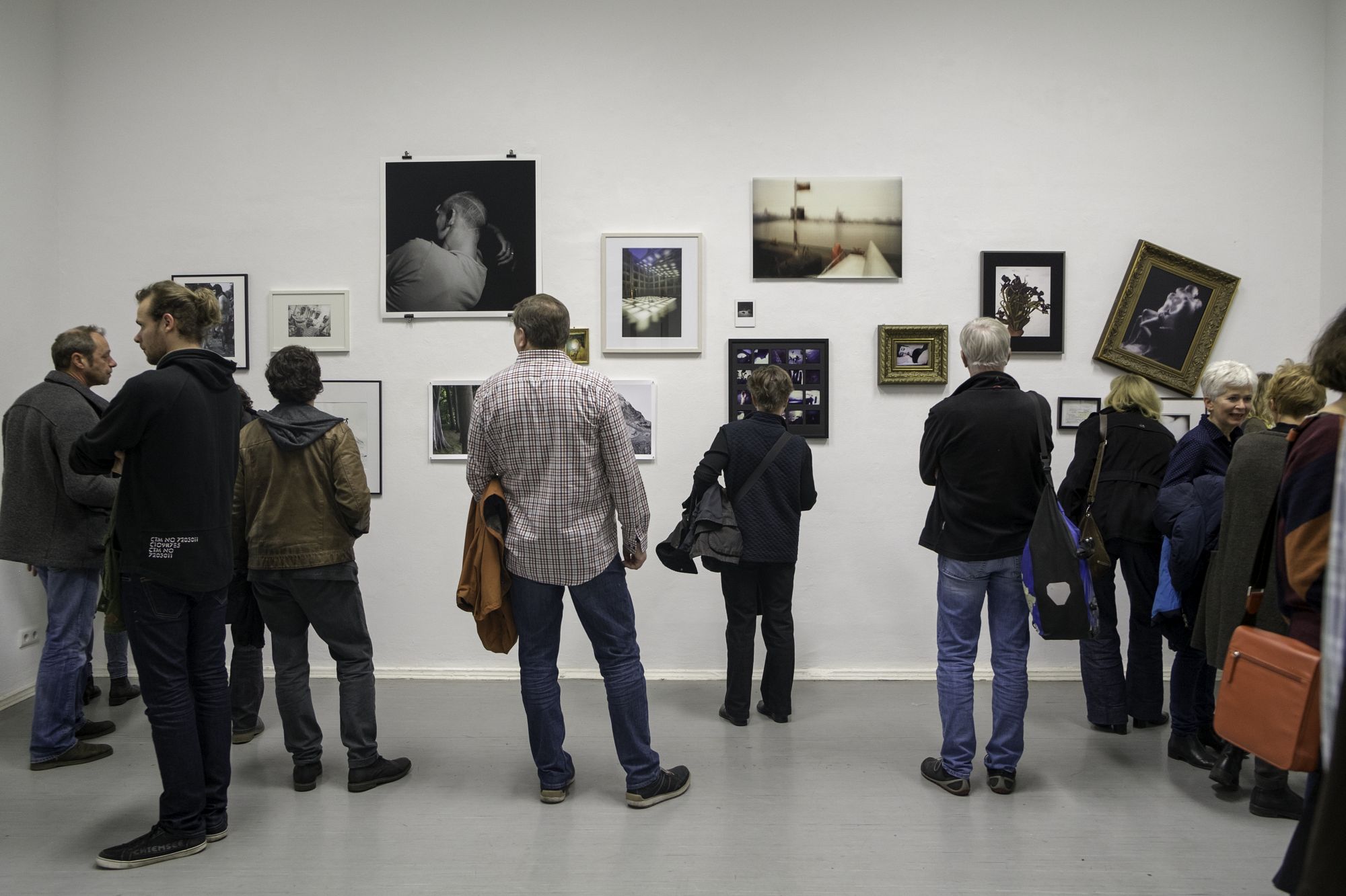 Menschen betrachten gerahmte Kunstwerke an einer weißen Wand in einer Galerie. (KI generierte Beschreibung)