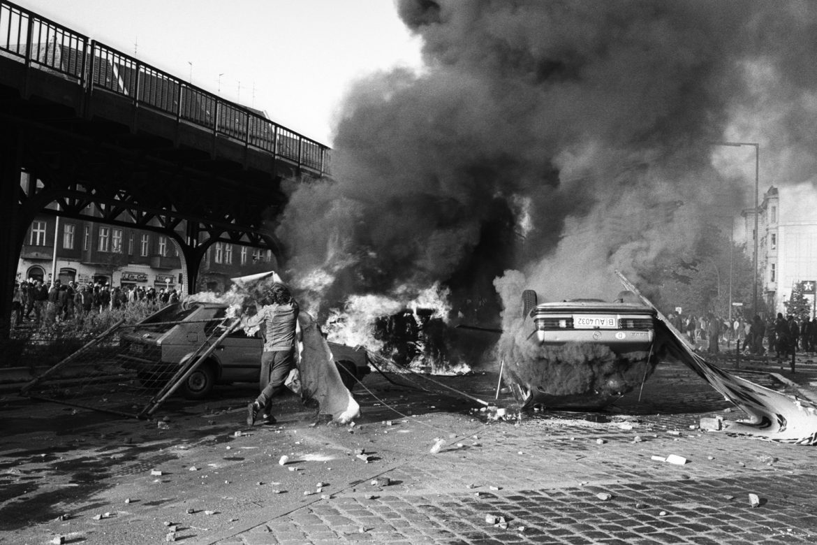 Eine Schwarz-Weiß-Szene ziviler Unruhen mit brennenden Autos und dichtem Rauch, auf einer Kopfsteinpflasterstraße verstreute Trümmer und Menschen im Hintergrund. (KI generierte Beschreibung)