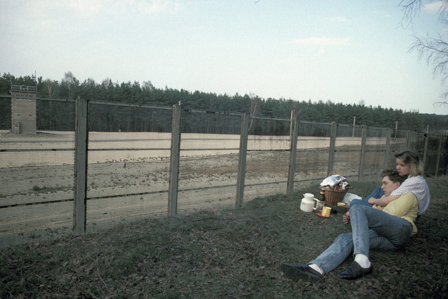 Zwei Personen sitzen mit einem Korb, einer Thermoskanne und Essen auf der Wiese neben einem Zaun. Sie wirken entspannt und blicken auf einen eingezäunten Bereich mit Bäumen im Hintergrund. (KI generierte Beschreibung)