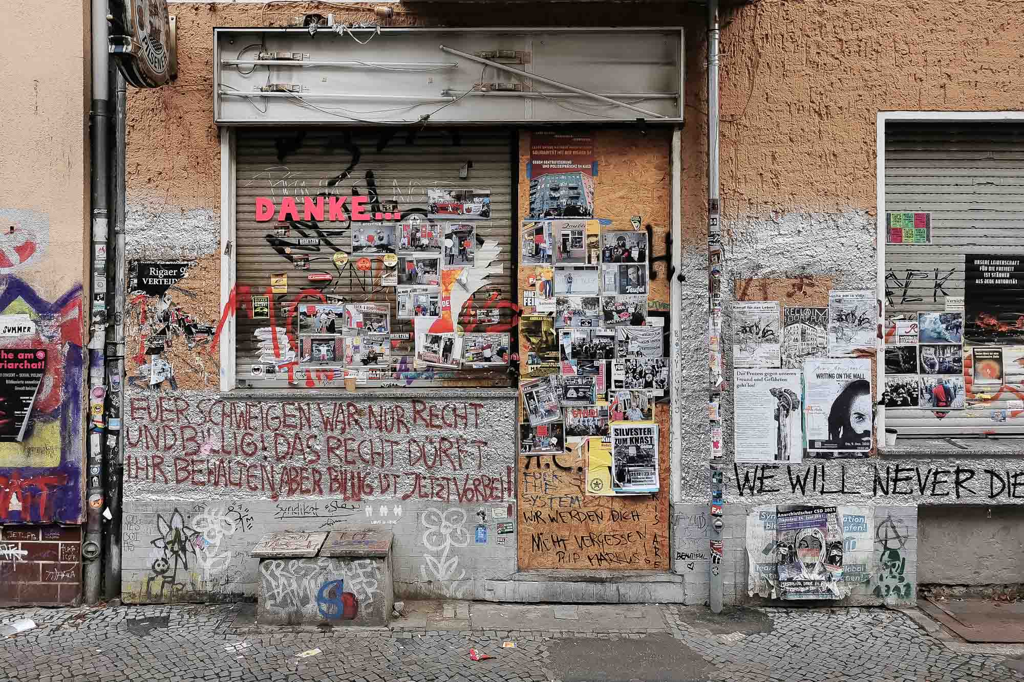Urbane Ladenfront mit bunten Plakaten, Graffiti und Aufklebern. Botschaften in verschiedenen Sprachen bedecken die Wände und Fensterläden. Der Bürgersteig säumt die reich verzierte Fassade. (KI generierte Beschreibung)