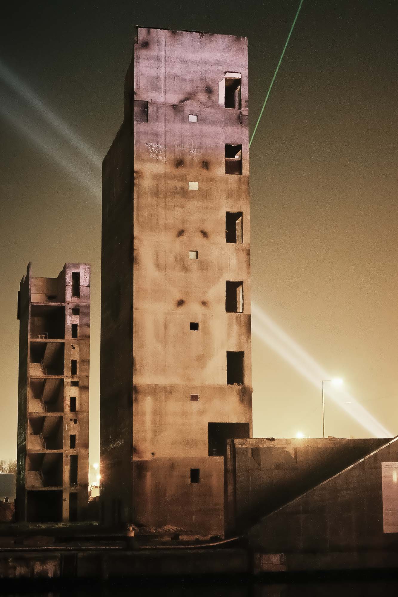 Zwei hohe, rechteckige, verlassene Betongebäude bei Nacht, mit teilweiser Beleuchtung und einem grünen Laserstrahl am Himmel. (KI generierte Beschreibung)