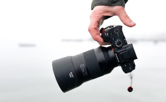 Eine Hand hält eine Sony-Digitalkamera mit einem großen Objektiv vor einem unscharfen, hellen Hintergrund. (KI generierte Beschreibung)