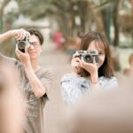 Zwei Personen halten Kameras in der Hand und fotografieren im Freien, dem Betrachter zugewandt, während eine weitere Person unscharf im Vordergrund steht. (KI generierte Beschreibung)
