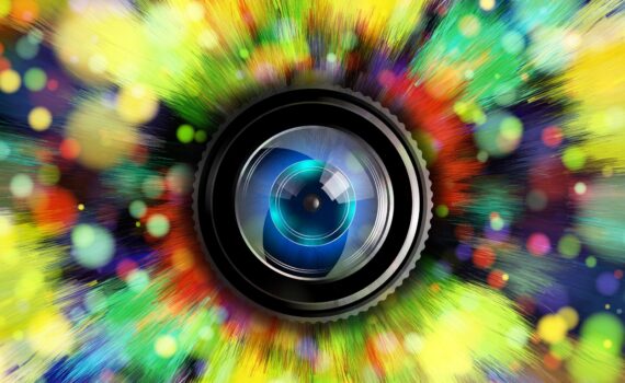 Eine Kameralinse steht in der Mitte eines lebhaften, farbenfrohen Lichtblitzes und eines Bokehs, wodurch ein Effekt entsteht, der an ein Auge erinnert. (KI generierte Beschreibung)