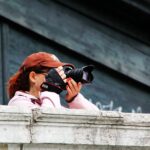 Eine Person mit brauner Mütze und rosafarbener Jacke steht hinter einem Steinsims, hält eine Kamera vor ihr Gesicht und macht ein Foto. (KI generierte Beschreibung)