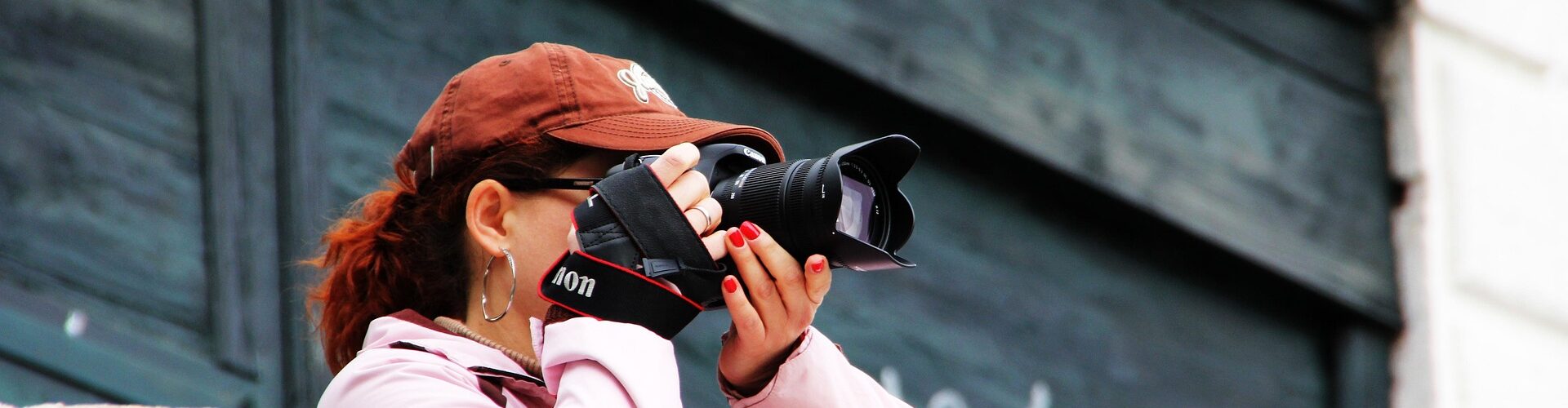 Eine Person mit brauner Mütze und rosafarbener Jacke steht hinter einem Steinsims, hält eine Kamera vor ihr Gesicht und macht ein Foto. (KI generierte Beschreibung)