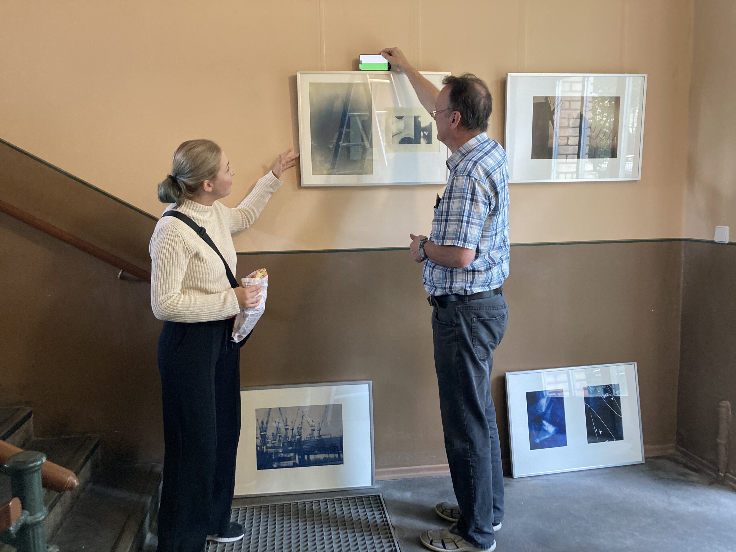 Ein Mann richtet ein gerahmtes Kunstwerk an der Wand aus, während eine Frau mit einer Snacktüte zuschaut; darunter lehnen weitere gerahmte Werke an der Wand. (KI generierte Beschreibung)