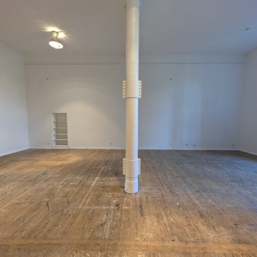 Geräumiger, leerer Raum mit Holzböden, weißen Wänden, einer großen, zentralen Säule, zwei eingebauten Wandregalen und einem Bogenfenster, das Tageslicht hereinlässt. (KI generierte Beschreibung)